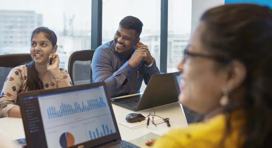 Colleagues sharing a joke around a meeting table. There are laptops with graphs visible on the table 