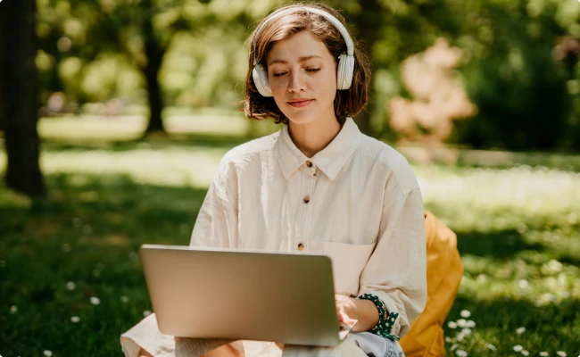 A woman sitting in the grass with a laptop wearing headphones