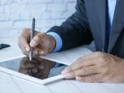 Person in a suit using a stylus on a tablet at a white desk.