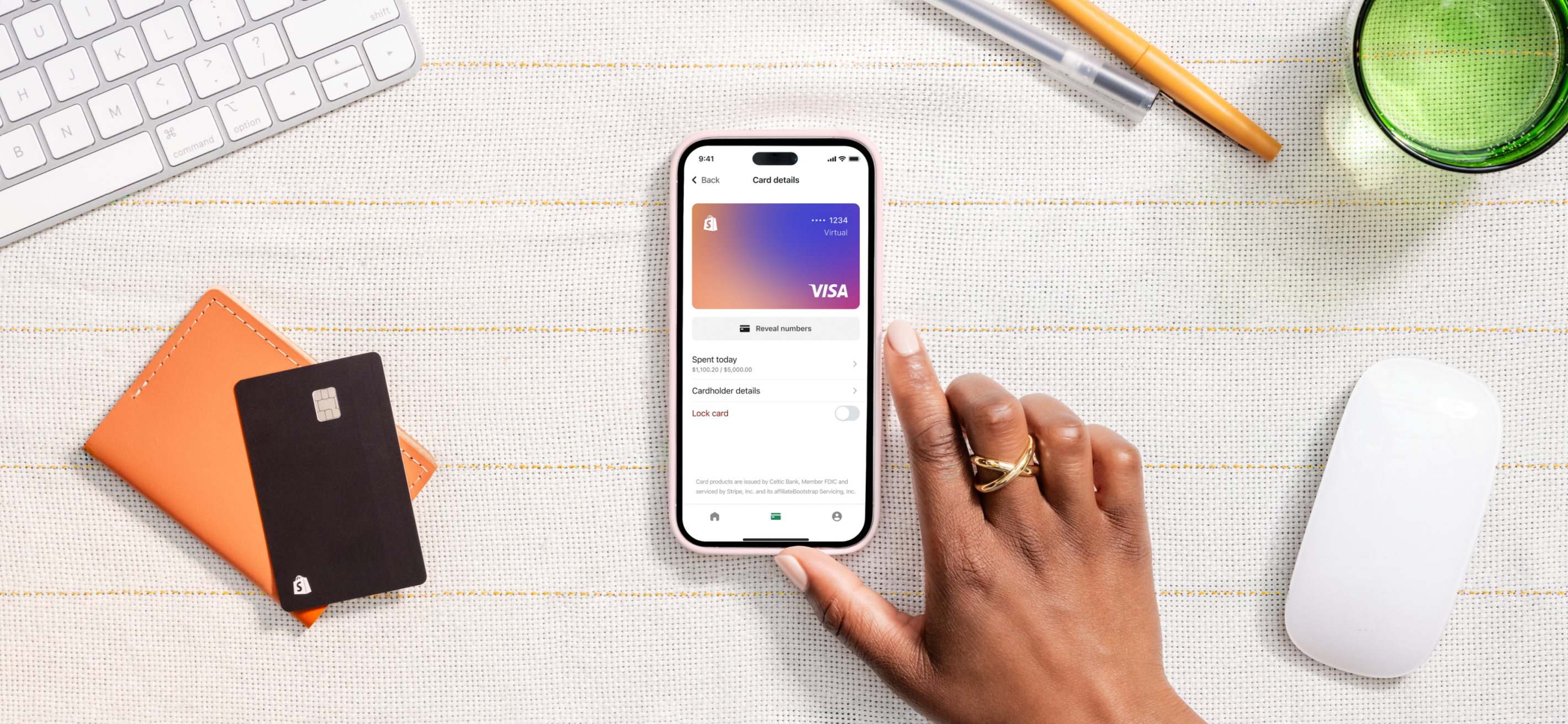 A merchant’s right hand touches a mobile device on a striped desk. The device displays the digital card of a Shopify Balance account.