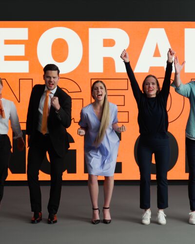 A group of people in various outfits is cheering together. In the background, there’s a board in SIXT colors with the word "Orange" written on it.