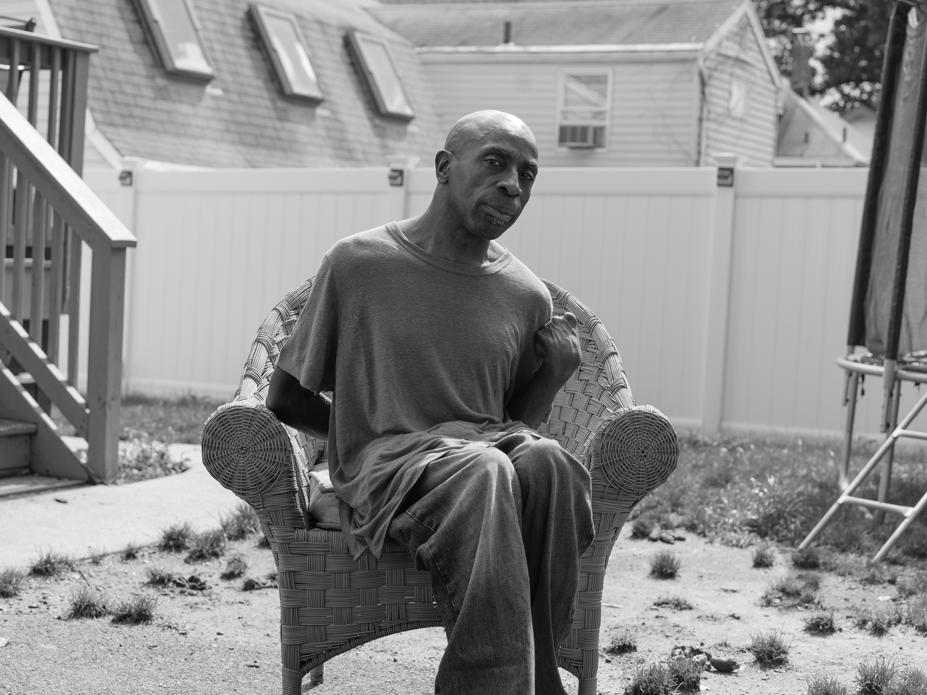 A black and white image of a man sitting in a wicker chair in the sun, in a yard sparsely covered with clumps of grass