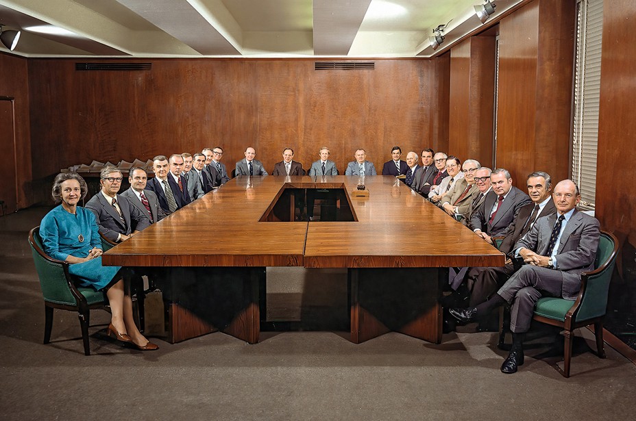 group photo of 23 people in suits, seated around large wooden conference table in wood-paneled conference room, with the woman in a turquoise suit