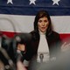 Photo of Nikki Haley speaking at a microphone, hands apart, with press before her and an American flag behind her