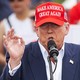 Wearing a red MAGA hat and a blue suit with a flag lapel pin, Trump speaks into a microphone at his recent rally in Las Vegas.