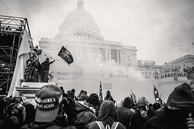 A photo of a crowd with a MAGA hat in the foreground and people standing on scaffolding with a Second Amendment flag and Trump banner in front of the smoke-obscured U.S. Capitol