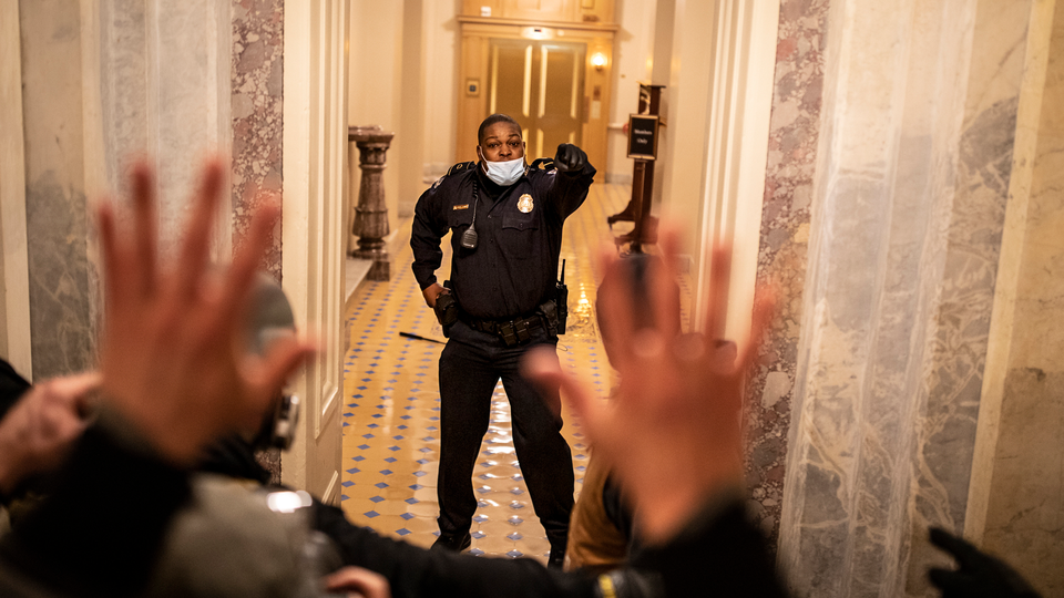 Photo of Capitol Police Officer Eugene Goodman standing in doorway of Capitol Building stairwell on January 6, 2021, with hand raised, opposite a crowd of rioters