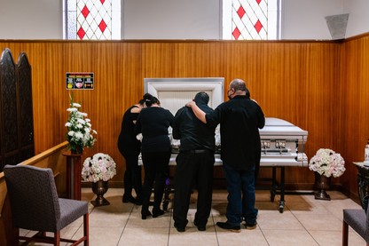 Family members dressed in black stand around a white casket, mourning at a funeral home.