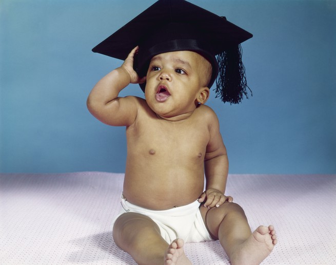 A baby clad in only a diaper holds a graduation cap on his head with an open-mouthed expression