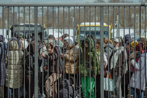 A line of people bundled up in parkas and scarves wait in a crowded line behind a tall barred metal fence with buses and snowy city in background