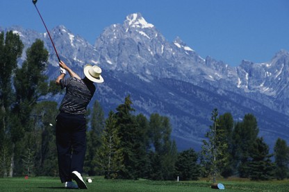 A golfer swings on a lush golf course with snow-capped mountains in the distance.