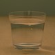 A short glass of water, resting on a light brown table, with an olive green wall in the background.