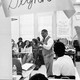 A black-and-white photo of a teacher teaching a class, captured through the window in the classroom door