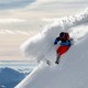 Photograph of a skier in red pants, a blue jacket, and a black helmet carving a turn in deep powder on a steep slope, with mountains in the background