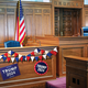 Photo-illustration of wood-paneled court chamber with judge's bench, American flag, and campaign bunting and posters saying "Trump 2024" around the witness stand