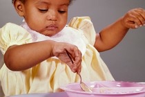 Color photo of a small child wearing a bib and eating off a pink plastic plate using a spoon