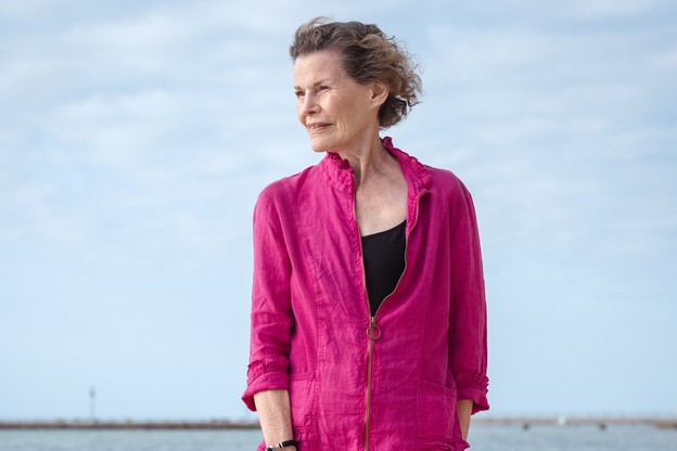 Woman in magenta top and black capri pants standing barefoot on beach with cloudy blue sky behind