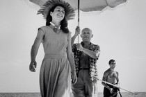 A photograph of Francoise Gilot walking on the beach ahead of Pablo Picasso, who is holding a sun umbrella