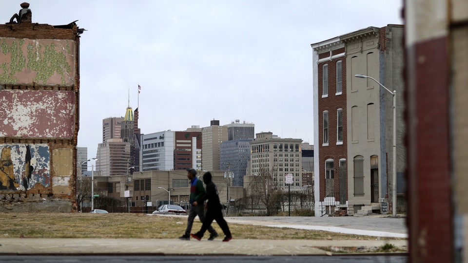 Two people walk past a vacant lot and boarded-up homes