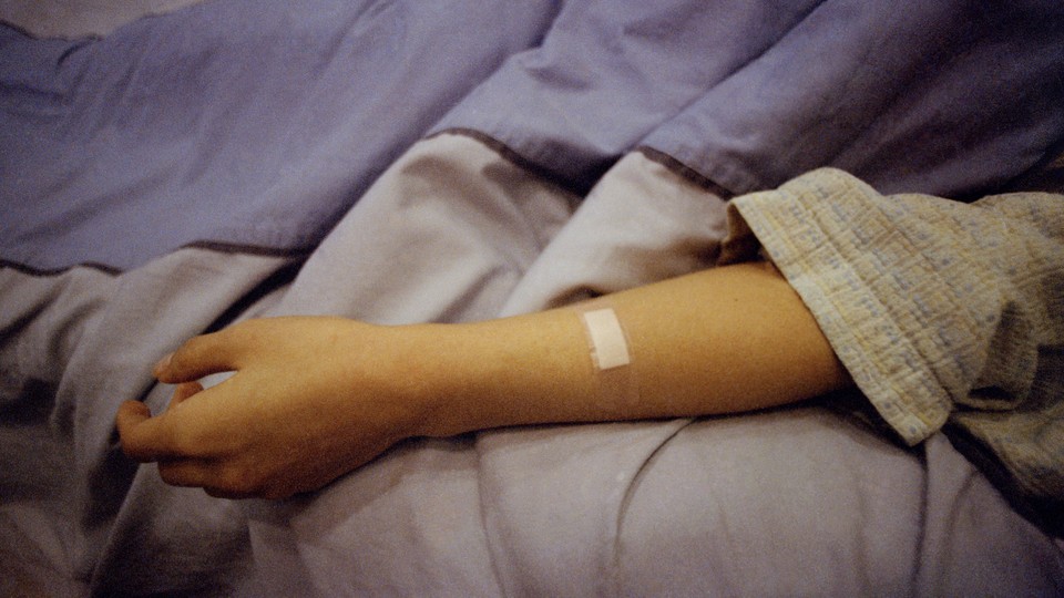 A patient's arm lies flopped on a bed, a bandage on the forearm