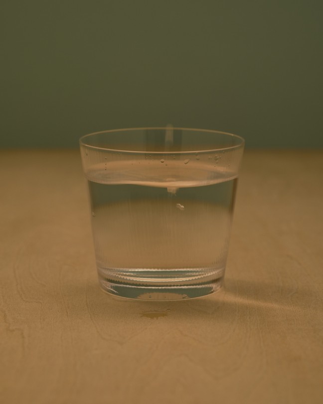 A short glass of water, resting on a light brown table, with an olive green wall in the background.