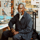 MR WASH sits in a chair facing the camera, with a solemn expression. The room around him is covered in artwork.