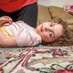 Girl with curly hair lying on colorful carpet with head turned toward the camera, with someone kneeling next to her and couch in background