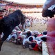 People lie on the ground in a small pile in front of a charging steer that jumps over them into a bullring full of other people.