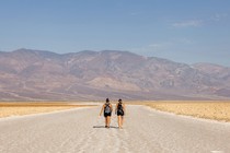 Two hikers walk on a path in front of barren mountains