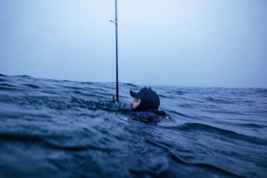 photo of man's head in hat and wetsuit just above an ocean swell, the rest of him submerged, with a large fishing pole vertical in water next to him