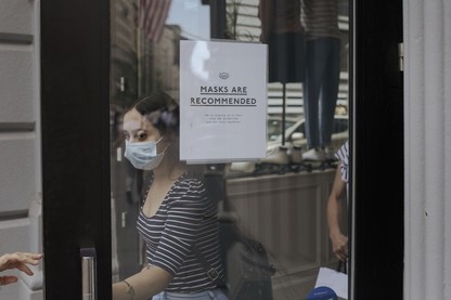 A woman wearing a mask opens a door. On the door is a sign that reads "Masks are recommended."