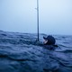 photo of man's head in hat and wetsuit just above an ocean swell, the rest of him submerged, with a large fishing pole vertical in water next to him