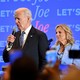 Joe Biden wearing a suit and speaking into a microphone in front of a backdrop reading "Let's Go Joe"; Jill is seated behind him.