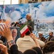 A white-haired man stands at a lectern, speaking to a crowd of people in front of a mural featuring his face.