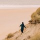 Photo of a person in a coat and hat scrambling along steep, grassy dunes next to a broad, sandy beach