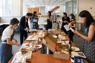 A long table filled with different gift baskets, surrounded by eight people sampling and discussing the contents.