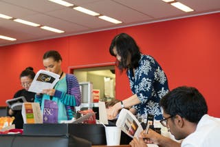 A group of people gathered around the gift basket sample table, in front of a bright red wall, seriously making notes on printed sheets.