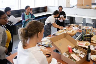 A long table filled with different gift baskets, surrounded by six people sampling and discussing the contents.