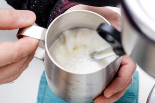 A person's hands holding a stainless steel pitcher of milk being steamed using the Bambino Plus espresso machine's wand.