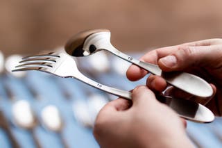 A tester holds up a spoon and fork, underside up, to show the maker's mark has been covered by a label that just says "19".