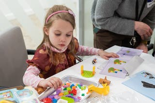 A young girl in a pink dress works on a toddler-friendly Lego kit.