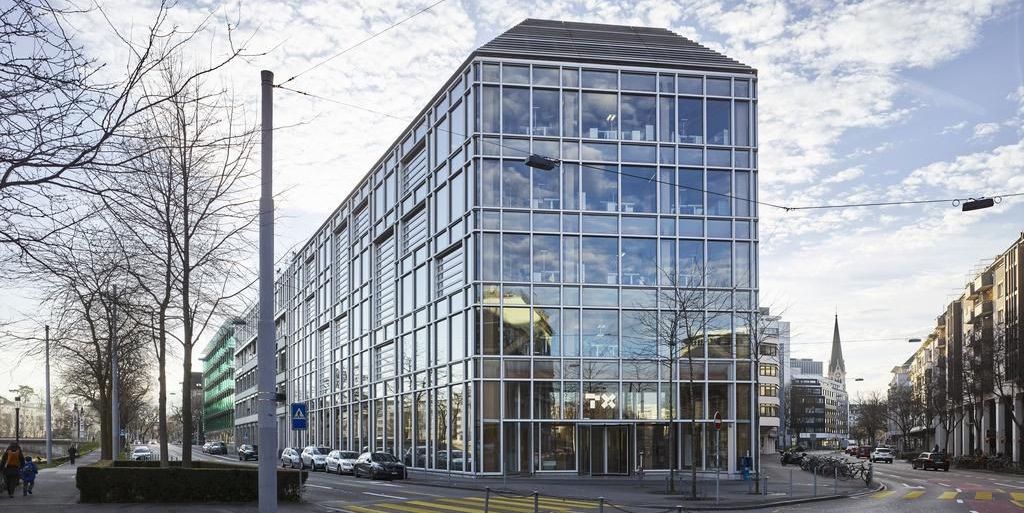 Moderne Glasgebäude an einer Strassenecke in einer Stadt, mit mehreren geparkten Autos entlang der Strasse und einem Kirchturm im Hintergrund unter einem bewölkten Himmel.