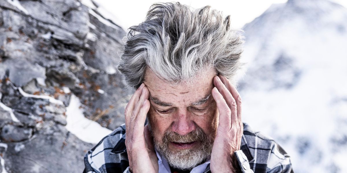 Älterer Mann mit grauem Haar steht im Freien vor einer schneebedeckten Berglandschaft und hält seinen Kopf mit beiden Händen.