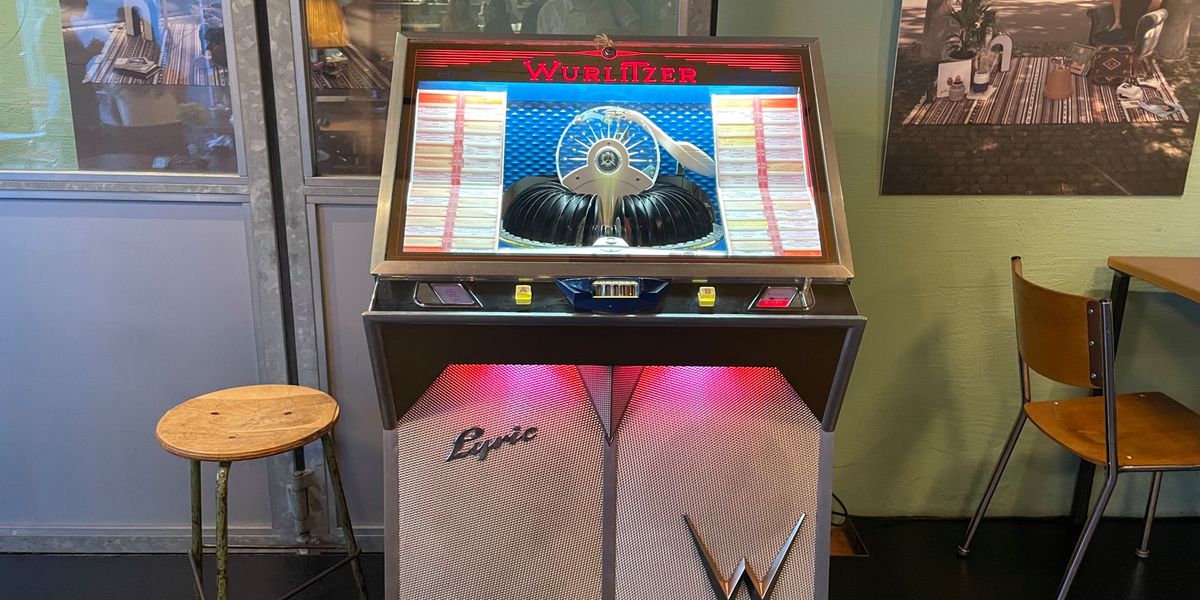 Eine klassische Wurlitzer-Musikbox steht in einem modernen Café neben einem runden Holzhocker und einem Tisch mit Stühlen.