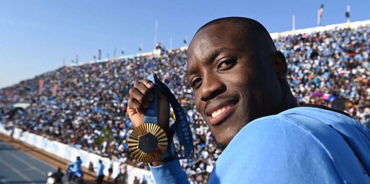 Letsile Tebogo hält seine olympische Goldmedaille und lächelt, während er in einem offenen Bus beim Willkommenszeremonie im Botswana National Stadium steht. Im Hintergrund jubelt eine grosse Menge von Menschen mit Botswana-Fahnen. 