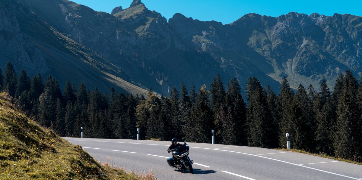 Un motard roulant sur une route de montagne entourée de forêts, avec des sommets en arrière-plan.