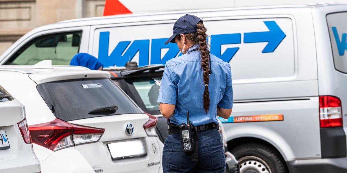 Eine Polizistin in Uniform schreibt ein Ticket für ein weisses Auto, das vor einem weissen Lieferwagen geparkt ist.