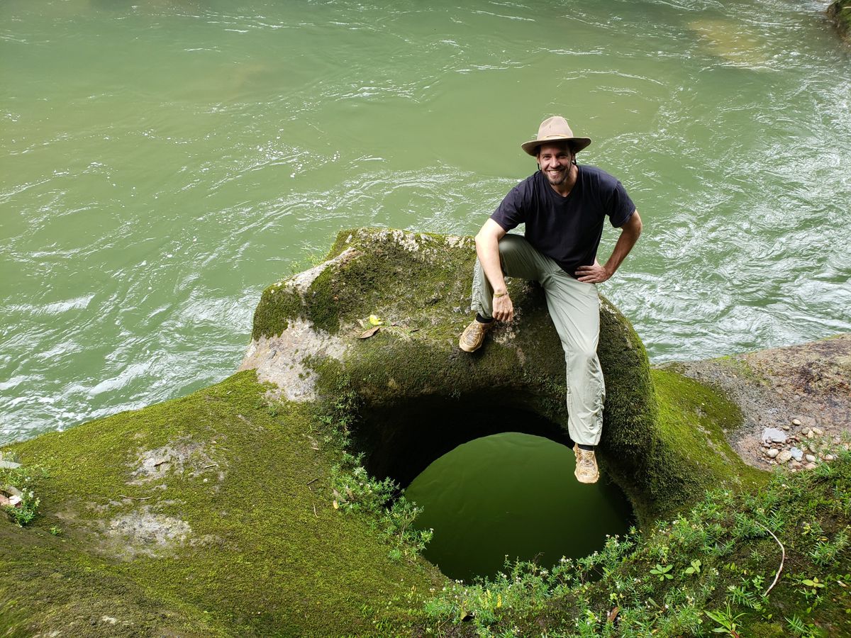 Ein Mann mit Hut sitzt entspannt auf einem moosbedeckten Felsen mit einem grossen Loch, daneben fliesst ein Fluss mit grünlichem Wasser.