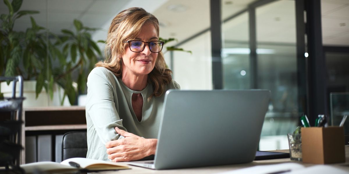 Senioren-Geschäftsfrau arbeitet im Büro an einem Laptop, liest Online-Projekte, Berichte und Recherchen. Kreativer Arbeitsplatz mit Pflanzen im Hintergrund.