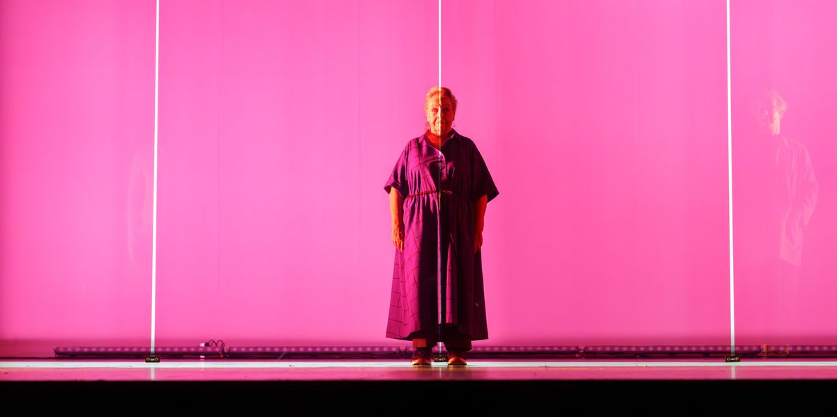 Ältere Frau in lila Kleid vor leuchtender rosa Wand stehend.